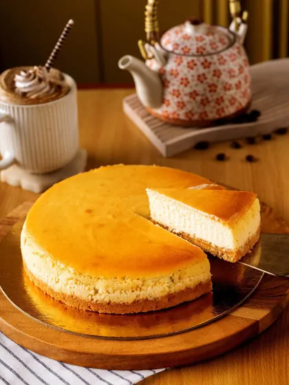 A classic New York cheesecake, with one slice removed to invite you in. The simple wooden board and accompanying coffee create a relaxed, cafe-style atmosphere, making the dessert feel accessible and delicious.