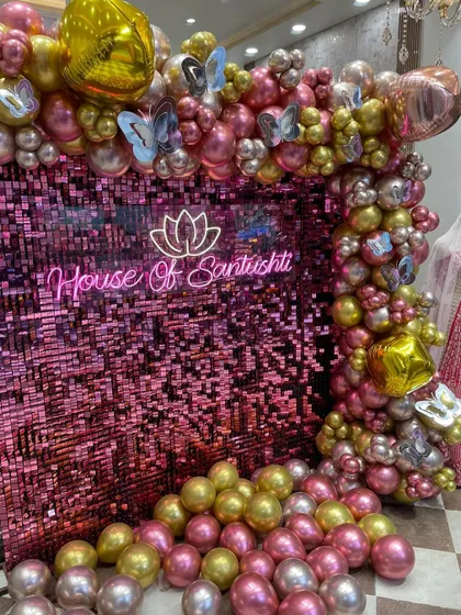 A glamorous setup for a store event, featuring a pink shimmer wall, a custom neon sign for "House Of Santushti," and a lush balloon frame in shades of pink, gold, and silver with butterfly accents.