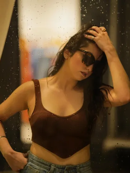 A dreamy, aesthetic shot through a rain-streaked window, wearing a brown crop top and sunglasses.