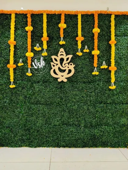 A simple and elegant greenery wall backdrop with a wooden Ganesha cutout. The wall is framed with an orange marigold border and features hanging floral tassels.