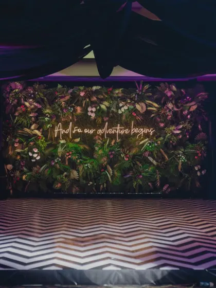 A lush, tropical photo wall with a neon sign reading "And so our adventure begins," perfect for a jungle-themed cocktail party.