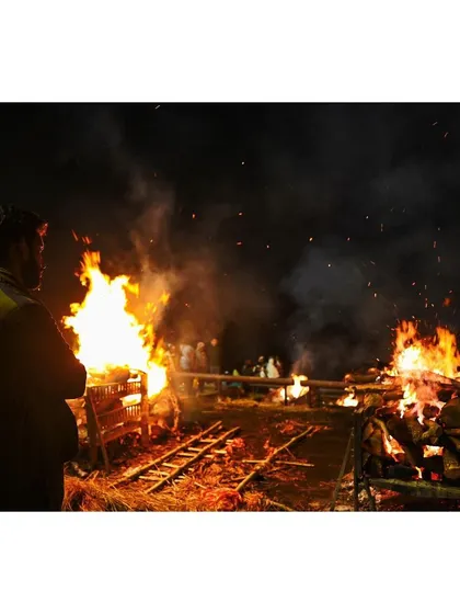 A man stands watching the intense flames of multiple funeral pyres at night. This image focuses on the raw power of the fire and the human element of observation and reflection.