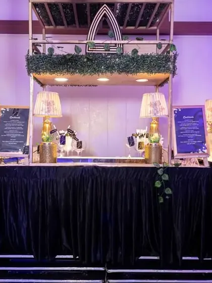 A stylish and modern bar setup for a cocktail party. The design features geometric accents, lush greenery, and elegant lamps, creating a sophisticated spot for guests to grab a drink.