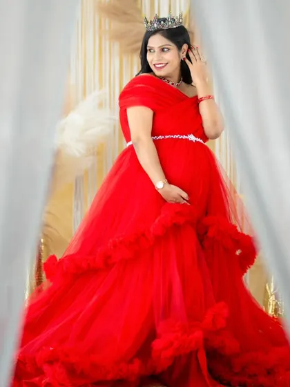 A candid moment of joy. The mom-to-be, wearing a vibrant red gown, looks off-camera with a beautiful smile, framed by the soft curtains of our studio.