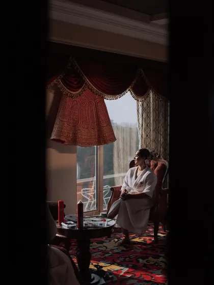 A creative shot of the bride getting ready. Her wedding lehenga hangs in the window, creating a sense of anticipation.