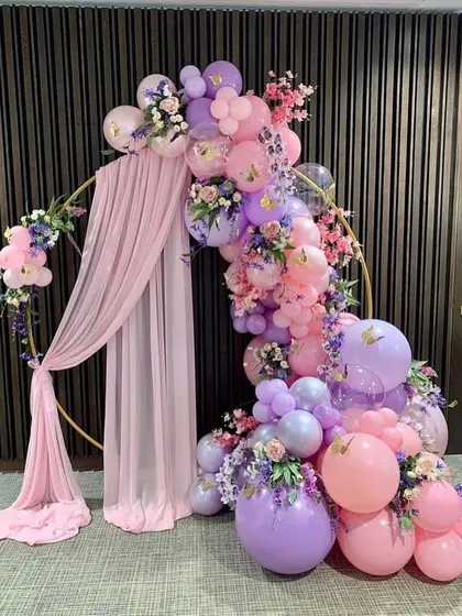 An elegant circular hoop decorated with pink and purple balloons, artificial flowers, and draped fabric, creating a sophisticated photo backdrop.