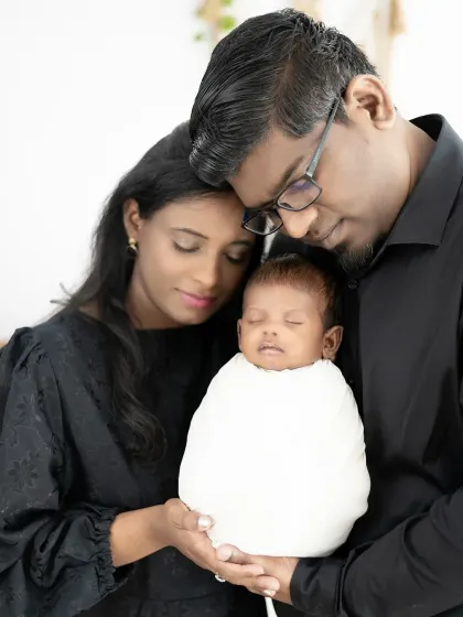 A beautiful, tight-knit portrait of new parents adoring their sleeping baby. The dark clothing and simple background make this an intimate and emotional image.