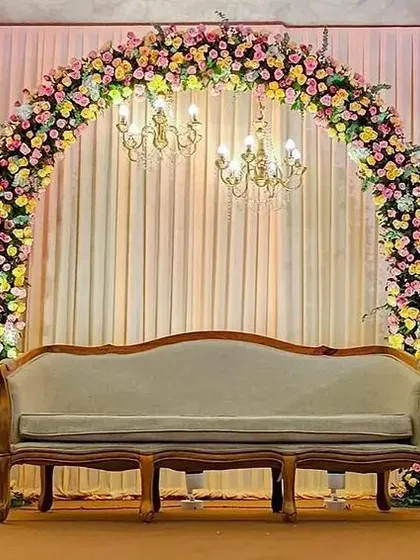 A beautiful stage setup featuring a full floral arch and multiple bouquets on tall, modern stands. The elegant chandelier adds a touch of sophistication to this engagement or reception decor.