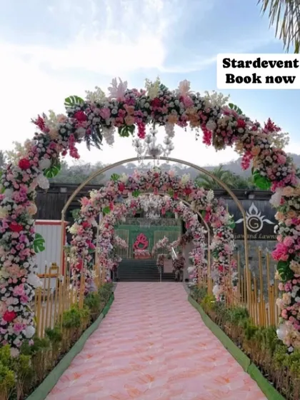 A beautiful daytime wedding entrance with lush floral arches in shades of pink and white. The pink patterned aisle and bamboo-style fencing create a charming and welcoming walkway.