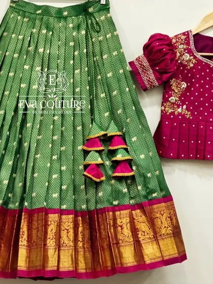 A beautiful green pattu pavadai with a contrasting magenta blouse. The blouse features delicate floral embroidery, and the green silk skirt has a traditional gold and red temple border.