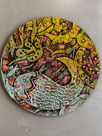 This finished circular collagraph artwork showcases a beautiful underwater scene with a fish. The artist used various textures and coloring techniques on the plate to achieve this rich, detailed effect.