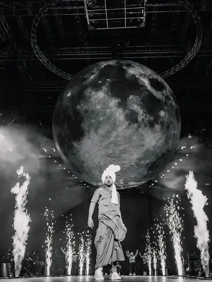 A highly artistic, black and white shot of Diljit Dosanjh on stage with a giant moon prop. This frame is about creating a larger-than-life, conceptual concert photograph.