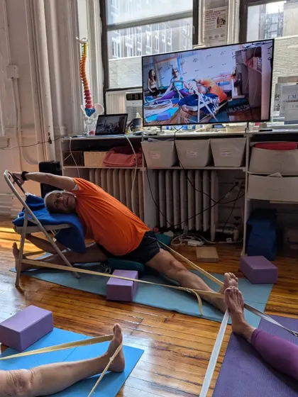 Teaching a hybrid class, I demonstrate a complex side stretch using a chair and straps. Students both in-person and online can follow the detailed alignment cues.
