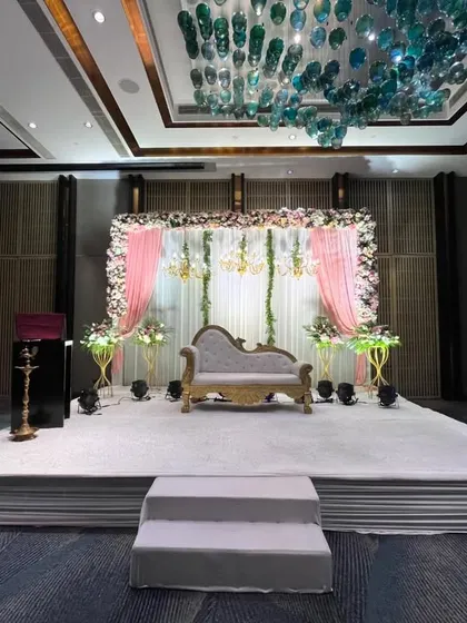 An elegant stage setup at Crowne Plaza Mayur Vihar. The design features a classic chaise lounge, pastel pink drapes, and hanging chandeliers under the hall's unique glass ceiling fixture.