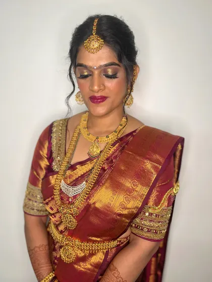 A stunning South Indian bridal look featuring golden shimmer on the eyes and a deep red lip. The makeup is perfectly balanced with the heavy gold jewelry and traditional silk saree.