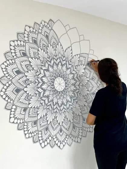 A work in progress shot of a large, grey mandala being painted directly onto a living room wall. Each layer is carefully added to build up the intricate, symmetrical design.