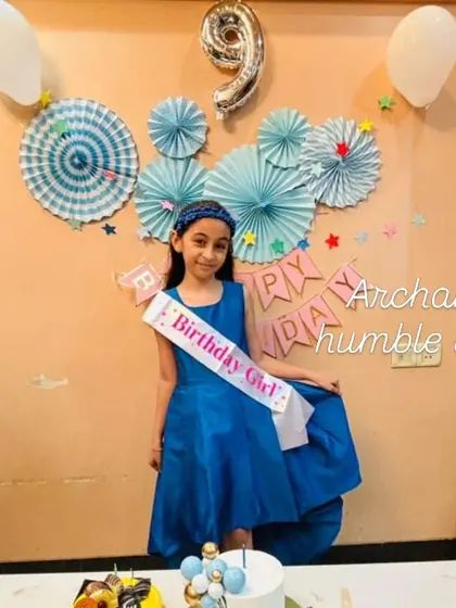 The birthday girl looking beautiful in her blue dress, standing by her balloon-themed cake.