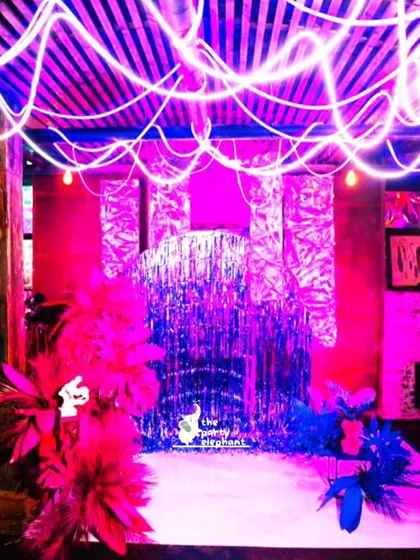 For this Ibiza-themed party, I created a vibrant photo booth with a silver fringe backdrop, dramatic pink and blue feather arrangements, and neon tube lighting for a high-energy, glamorous look.