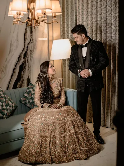 A full-length portrait showcasing the couple's impeccable style. The bride, seated in her magnificent golden lehenga, looks up at her groom, who stands beside her in a sharp tuxedo, creating a look of regal elegance.
