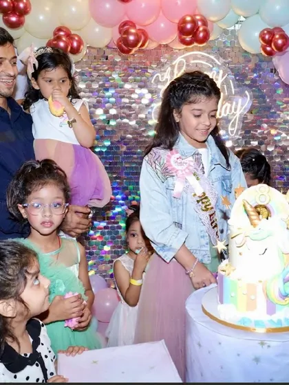 The birthday girl, wearing a "Birthday Girl" sash, cuts her multi-layered unicorn cake surrounded by her friends.