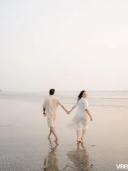 A couple walks along the wet sand, holding hands and looking back at the camera. The reflection in the sand and the soft light make this a beautiful, serene pre-wedding photograph.