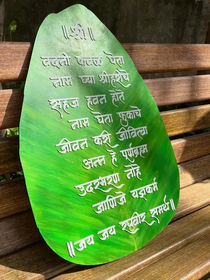 The leaf-shaped 'Vadani Kaval' wall hanging resting on a bench, showing its beautiful texture and the grace of the Marathi calligraphy.