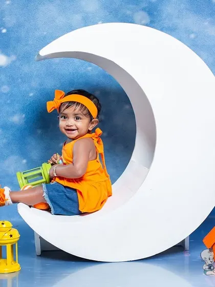 A happy toddler sitting on a large crescent moon prop against a starry night sky. The colorful paper lanterns add a festive and whimsical touch to this celestial-themed photo.