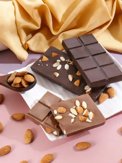 This chocolate photography uses soft, warm lighting and flowing fabrics to create a gentle, indulgent mood. The composition highlights different types of almond chocolate, showcasing the brand's variety.