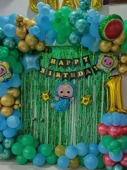 A Cocomelon-themed first birthday decoration for a home party. It features a green shimmer curtain, a balloon arch, Cocomelon character foil balloons, and a number '1' balloon pillar.
