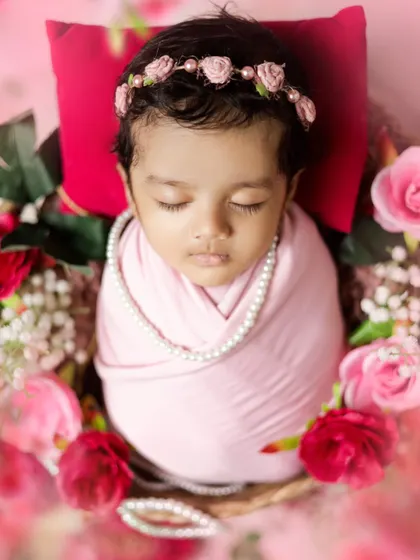 Pretty in pink. This newborn girl is wrapped snugly and adorned with a pearl necklace and a floral headband, surrounded by soft roses.