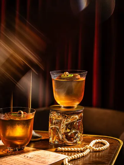 A dramatic, low-light photo of cocktails at Baglami. The use of light rays and shadows creates a mysterious, cinematic feel, perfect for a speakeasy-style bar.