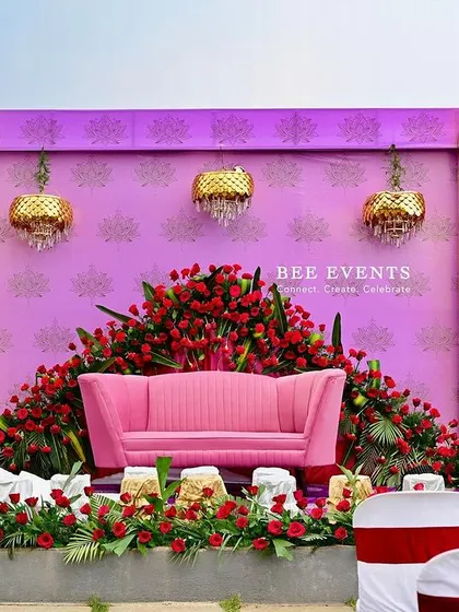 A vibrant stage setup featuring a pink sofa enveloped in a cascade of red roses. The design is framed by golden arches with red draping, creating a look that is both romantic and regal.