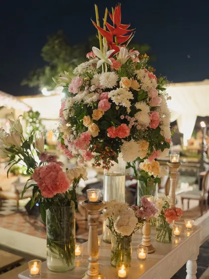 A close-up of a large floral centerpiece combining pink, white, and orange flowers with tropical leaves, surrounded by candles for a warm, inviting glow.