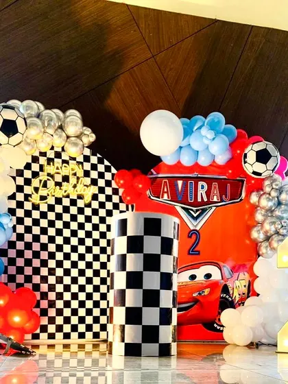 A close-up of a Cars-themed second birthday party. The decor includes a checkered flag backdrop, a Lightning McQueen cutout, and a marquee number '2'.