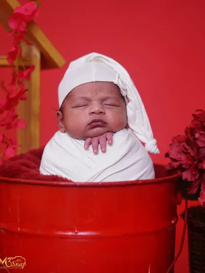 A newborn wrapped in white, resting in a red bucket with a sleepy hat. The bold red color creates a striking and memorable portrait.