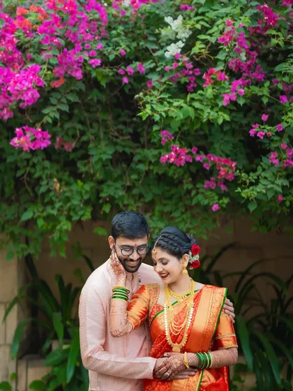A beautiful outdoor portrait of a Maharashtrian couple, with the groom gently touching the bride's face, framed by vibrant bougainvillea flowers.