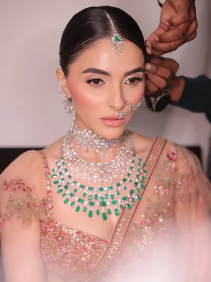 A behind-the-scenes moment as the final touches are made to the hair. Her makeup is soft and luminous, with a pop of pink on the cheeks to give her that fresh, bridal glow.