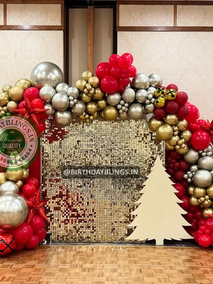 A festive Christmas-themed celebration with a gold shimmer wall and a balloon arch in red, gold, and silver. The Christmas tree cutout adds a perfect seasonal touch.