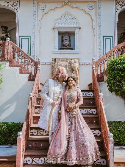 A tender moment as the groom kisses his bride's forehead on the steps of The Raj Palace in Jaipur, surrounded by intricate architecture.