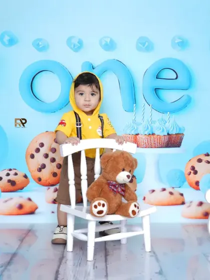 A baby boy stands proudly in his cookie-themed first birthday setup. The giant cookies and blue balloons create a playful and celebratory atmosphere.