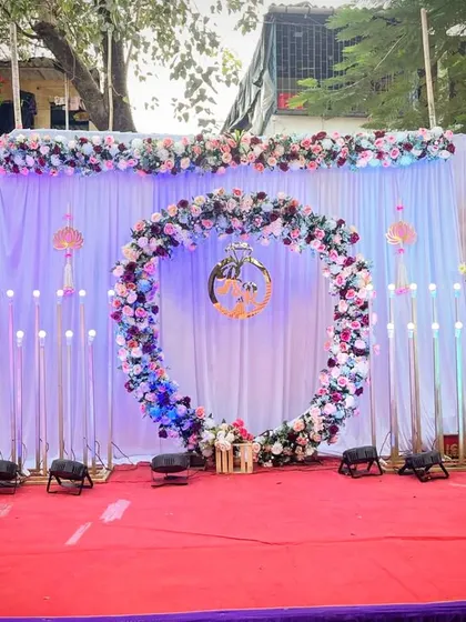 A lovely outdoor engagement setup on a red carpet. The centerpiece is a large floral ring decorated with pink and white flowers, set against a white curtain backdrop and flanked by modern, tall candle-like lights.