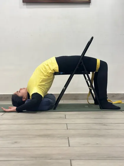 This is Chatushpadasana (Four-Footed Pose) performed with the support of a chair and belt. This setup helps build strength in the back and legs while ensuring proper alignment, making it a safe way to prepare for deeper backbends.