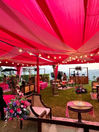 A vibrant outdoor Mehendi or Sangeet setup under a bright pink tent. The space is filled with comfortable lounge seating and colorful decor, perfect for a daytime celebration.