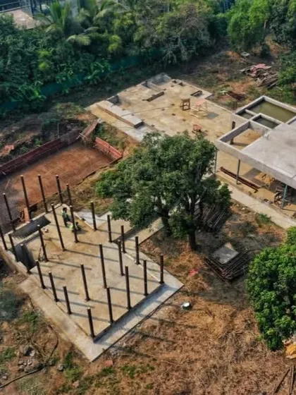 An aerial drone shot showing the foundation and plinth work for the Awas House. The layout is carefully planned to navigate the existing trees and natural contours of the land.