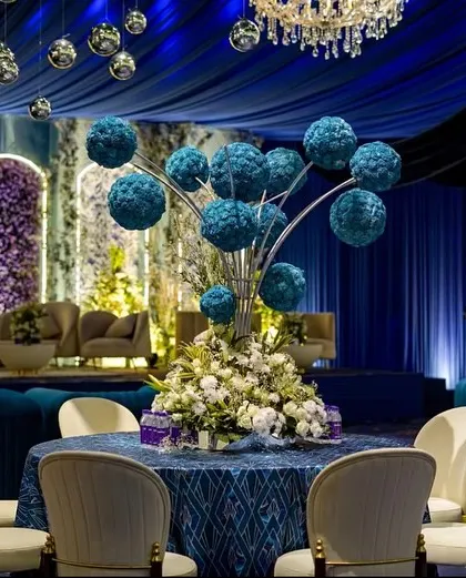 A table centerpiece featuring a modern arrangement of blue floral spheres. The backdrop shows the stage area with its beautiful arched panels and floral walls.