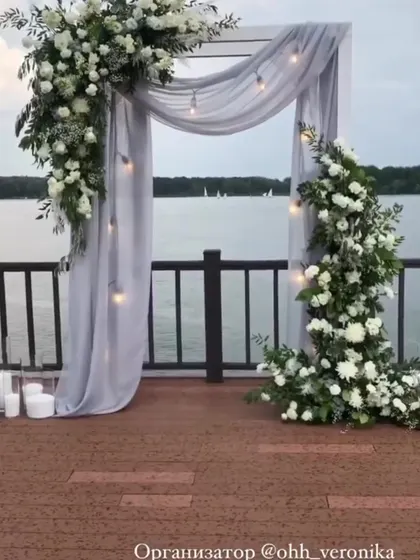A romantic photo spot by the water, with a simple frame draped in grey fabric and adorned with white floral arrangements.