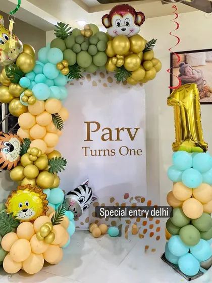 A first birthday jungle theme setup featuring a custom backdrop for "Parv Turns One". The decor includes a balloon arch with animal foil balloons and a matching balloon pillar with a large number '1' foil balloon.