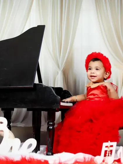 A one-year-old girl in a festive red dress plays the piano to celebrate her birthday.