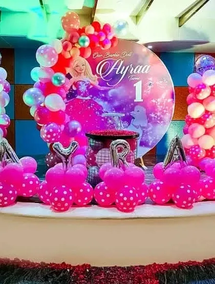 A Barbie-themed first birthday party on a stage. This decoration features a "Barbie Doll" backdrop, balloon columns, and hot pink balloon arrangements.