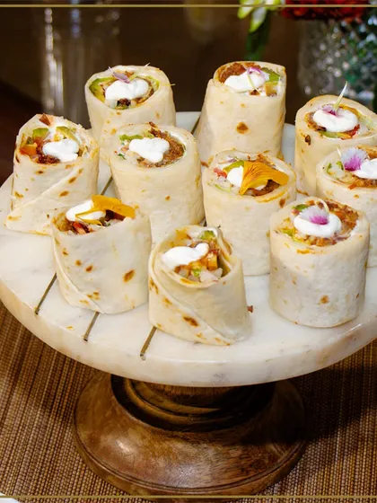 Bite-sized tortilla wraps filled with fresh ingredients and a dollop of cream, presented on a marble and wood cake stand. These are easy for guests to enjoy while mingling.